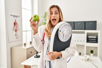 Sticker - Young blonde doctor woman holding weighing machine and green apple celebrating crazy and amazed for success with open eyes screaming excited.