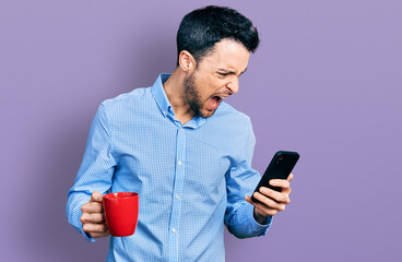 Poster - Hispanic man with beard using smartphone and drinking a cup of coffee angry and mad screaming frustrated and furious, shouting with anger. rage and aggressive concept.