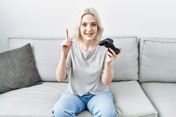 Sticker - Young caucasian woman at home playing video game holding controller smiling with an idea or question pointing finger with happy face, number one