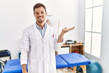 Wall Mural - Handsome young man working at pain recovery clinic smiling cheerful presenting and pointing with palm of hand looking at the camera.