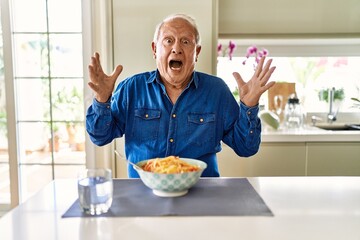 Canvas Print - Senior man with grey hair eating pasta spaghetti at home celebrating crazy and amazed for success with arms raised and open eyes screaming excited. winner concept