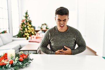 Canvas Print - Young hispanic man sitting on the table by christmas tree with hand on stomach because indigestion, painful illness feeling unwell. ache concept.