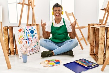 Sticker - Young hispanic man at art studio shouting with crazy expression doing rock symbol with hands up. music star. heavy concept.