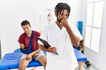 Poster - Young hispanic man working at pain recovery clinic with a man with broken arm smelling something stinky and disgusting, intolerable smell, holding breath with fingers on nose. bad smell