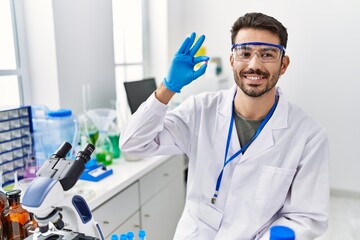 Wall Mural - Young hispanic man wearing scientist uniform holding pill at laboratory