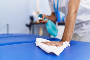 Poster - Young hispanic man wearing physiotherapist uniform disinfecting massage table at rehab clinic