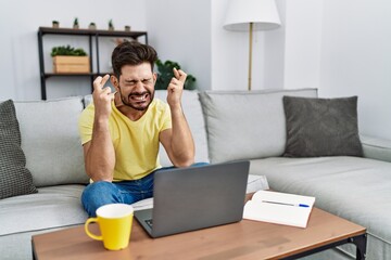 Poster - Young man with beard using laptop at home gesturing finger crossed smiling with hope and eyes closed. luck and superstitious concept.