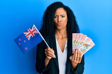 Sticker - Middle age african american woman holding new zealand flag and dollars making fish face with mouth and squinting eyes, crazy and comical.