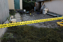 Victim Of A Violent Crime In A Backyard Of Residental House In Evening. Dead Man Body Under The Yellow Police Line Tape And Evidence Markers On Crime Scene