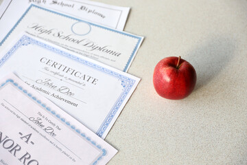 A honor roll recognition, certificate of achievement and high school diploma lies on table with red apple. Education documents