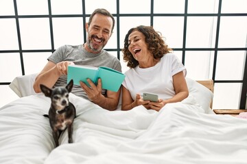 Sticker - Middle age hispanic couple reading book and using smartphone. Lying on the bed with dog at home.