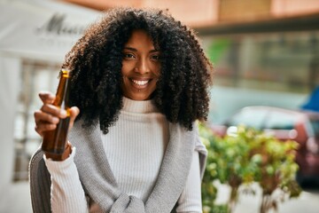 Poster - Young african american woman smiling happy holding bottle of beer at the city.