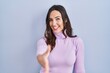 © Krakenimages.com - Young brunette woman standing over blue background smiling friendly offering handshake as greeting and welcoming. successful business.