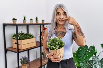 Sticker - Middle age grey-haired woman holding green plant pot at home pointing with hand finger to face and nose, smiling cheerful. beauty concept