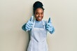 African american woman with braided hair wearing cleaner apron and gloves approving doing positive gesture with hand, thumbs up smiling and happy for success. winner gesture.