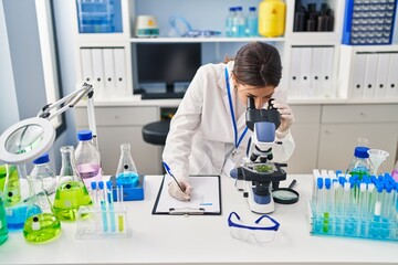 Canvas Print - Young hispanic woman wearing scientist uniform using microscope and writing on clipboard at laboratory