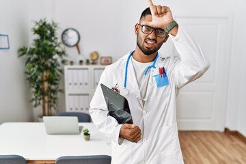 Canvas Print - Young indian man wearing doctor uniform and stethoscope making fun of people with fingers on forehead doing loser gesture mocking and insulting.