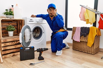 Canvas Print - Young indian technician working on washing machine peeking in shock covering face and eyes with hand, looking through fingers with embarrassed expression.