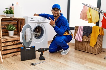 Canvas Print - Young indian technician working on washing machine cheerful with a smile on face pointing with hand and finger up to the side with happy and natural expression