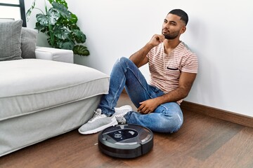 Wall Mural - Young indian man sitting at home by vacuum robot serious face thinking about question with hand on chin, thoughtful about confusing idea