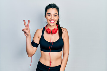 Young hispanic girl wearing gym clothes and using headphones showing and pointing up with fingers number two while smiling confident and happy.