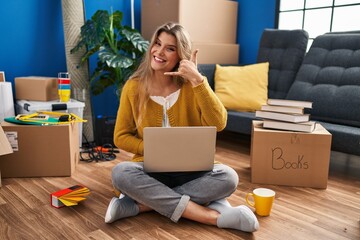 Sticker - Young woman sitting on the floor at new home using laptop smiling doing phone gesture with hand and fingers like talking on the telephone. communicating concepts.