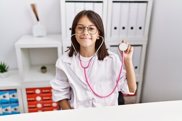 Poster - Young hispanic girl wearing doctor coat holding stethoscope looking positive and happy standing and smiling with a confident smile showing teeth