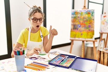 Sticker - Young brunette teenager at art studio pointing down with fingers showing advertisement, surprised face and open mouth