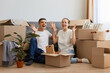 © sementsova321 - Full length portrait of joyful newlyweds moved in new apartment, sitting on the floor among cardboard boxes, raised hands, rejoicing to rent a house together.