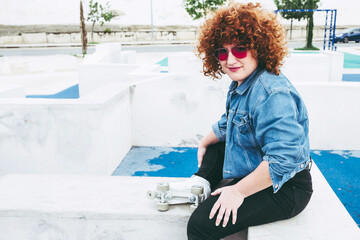 Young curly redhead woman with roller skates. Body positive girl enjoys in the park doing sports. Miss with vintage style and colored sunglasses. Portrait smiling in urban landscape. Large space copy