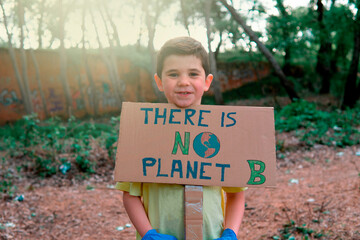 Young boy wearing a yellow t-shirt and blue gloves holding a cardboard sign drawn by hand with the symbol of a terrestrial globe and on which he puts an ecological awareness message.