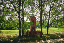 A Red Brick Wayside Shrine Standing By A Dirt Road In The Shade Of Trees. Tuchola Forest, Poland