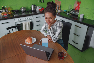 Wall Mural - mixed-race young beautiful female sits in kitchen at table looking at laptop, video call
