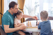 © Elenka - A cute blond boy and a girl play chess with their father, have fun together at home. A man teaches children to play a game, play a board game, play chess. Family times.