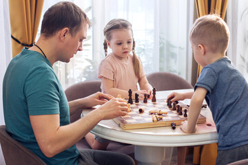 A cute blond boy and a girl play chess with their father, have fun together at home. A man teaches children to play a game, play a board game, play chess. Family times.