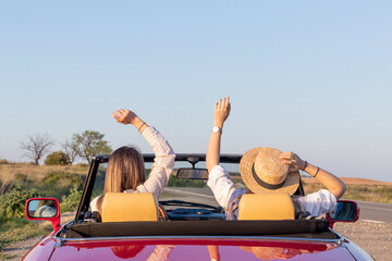 Happy free girlfriends with hat driving in red retro car cheering joyful with arms raised. Road trip travel concept