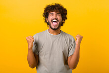 Overjoyed Crazy Happy Indian Curly Guy Screaming Yes In Ecstatic, Raising Fists Up, Celebrating Good Luck, Victory, Isolated On Yellow. Goal Achievement Concept