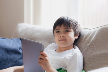 Wall Mural - Cinematic portrait young boy playing game on tablet sitting on sofa with light shining from window, Kid playing games online on internet at home, Child talking video call with friends at home