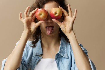 Wall Mural - Funny cute curly female in jeans casual shirt closes eyes with apples sticks tongue out posing isolated on over beige pastel background. Healthy food. Natural eco-friendly products concept. Copy space