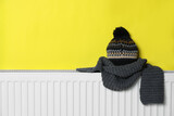 Fototapeta Kawa jest smaczna - Knitted hat and scarf on heating radiator near yellow wall, space for text