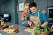 Happy lesbian drinks coffee and embraces her girlfriend who is preparing food in kitchen.