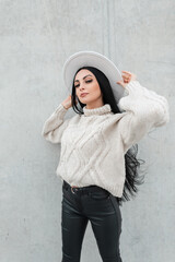 Wall Mural - Fashion pretty woman with glasses in casual urban clothes with white knit sweater, leather pants and white hat poses near a gray wall on the street