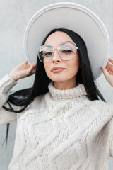 Wall Mural - Beautiful young stylish woman with glasses in a vintage fashionable sweater and hat walking in the city