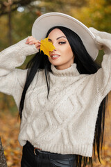 Wall Mural - Happy beautiful young woman model with smile in white knitted sweater and hat holds a autumn yellow leaf and walks in fall colorful park