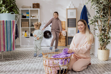 Wall Mural - Little girls are playing in the bathroom, laundry room, dancing, running while mom is busy doing chores, folding clothes, preparing things to put in the washing machine.