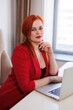 © Tatyana - Business woman sitting at her table in office. Red-haired girl manager working in computer. Office worker in red suit in her gorgeous cabinet. Company director sitting in armchair, book-case behind