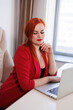 © Tatyana - Business woman sitting at her table in office. Red-haired girl manager working in computer. Office worker in red suit in her gorgeous cabinet. Company director sitting in armchair, book-case behind