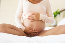 Mid Section Of Caucasian Pregnant Woman Touching Her Belly While Sitting On The Bed At Home