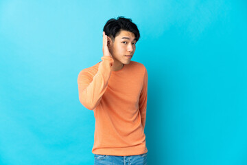 Wall Mural - Young Chinese man isolated on blue background listening to something by putting hand on the ear