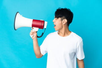 Young Chinese man isolated on blue background shouting through a megaphone to announce something in lateral position